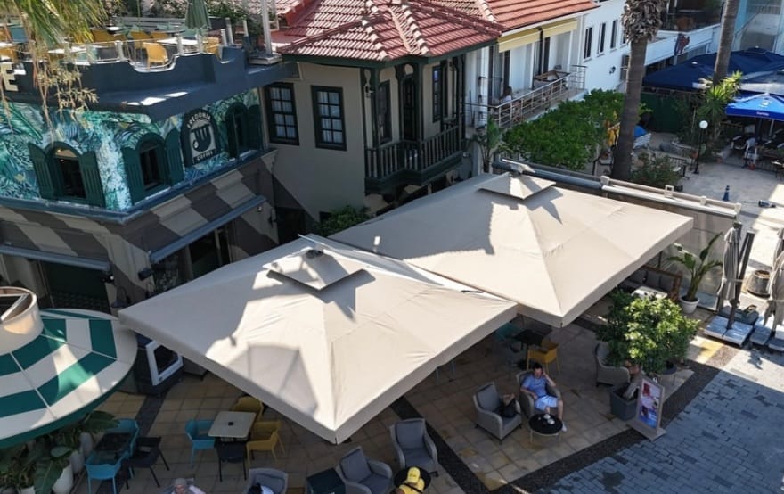 Polo Bilgi Bankası; ticari açık alanlarda doğru ölçü, ürün adedi, yerleşim, sabitleme ve güvenli kullanım kararlarını teknik rehberlerle destekler. Cafe ve restaurant açık alanında yan yana konumlandırılmış krem kumaşlı iki büyük teleskopik şemsiye uygulaması